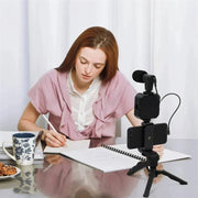 Woman using smartphone vlogging kit with tripod, microphone, and LED fill light for video recording at a desk